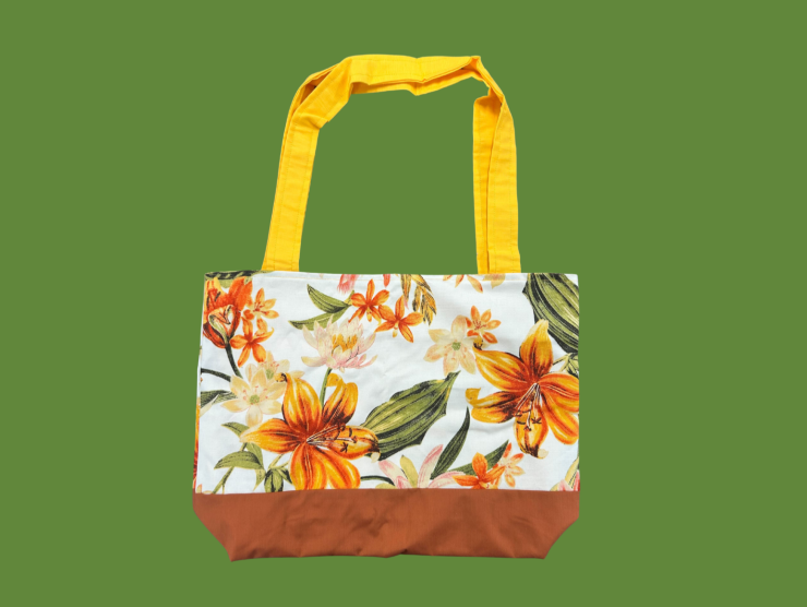 A green background with a wide totebag that has yellow handles, a tropical floral print fabric body and a brown bottom.