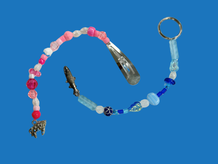 Blue background with a string of assorted pink and white beads connected to a barrette on one end and a silver cat charm on the other, and a blue string of assorted blue and white beads connected to a key chain ring on one end and silver fish charm on the other.