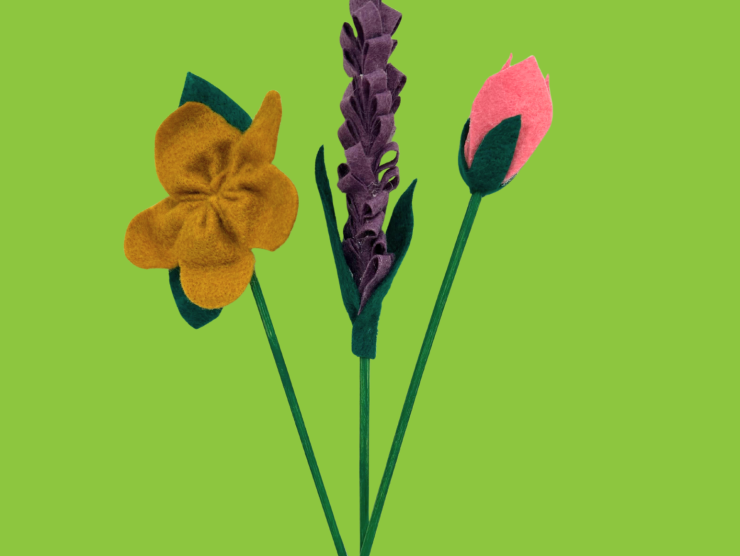 A bright green background with three felt flowers, a five petal yellow flower, a purple lavendar style flower and a pink tulip style flower.